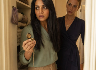 A woman clutches an open locket in a linen closet as a blonde woman with a scarred eyebrow watches from the doorway.