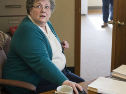 A gray-haired woman in glasses freezes at her desk as someone stops in her doorway.