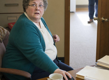 A gray-haired woman in glasses freezes at her desk as someone stops in her doorway.