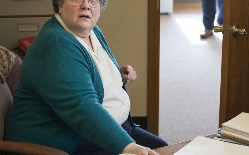 A gray-haired woman in glasses freezes at her desk as someone stops in her doorway.