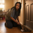 A Black woman with long box braids kneels by her front door, lifting the doormat to reveal a paper slipped underneath.