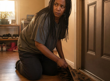A Black woman with long box braids kneels by her front door, lifting the doormat to reveal a paper slipped underneath.